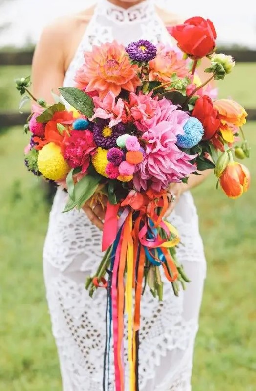 a colorful boho wedding bouquet with red, pink and purple blooms, foliage, colorful pompoms and long colorful ribbons