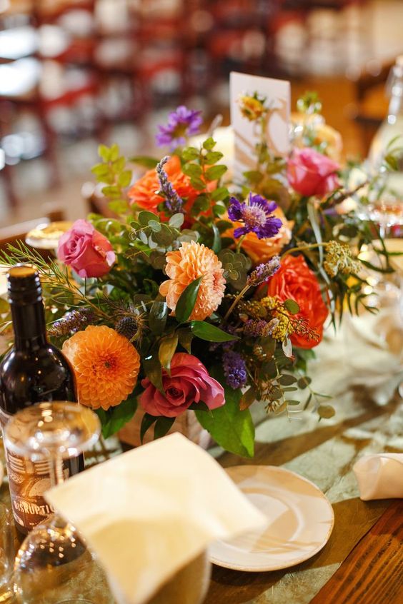 a colorful wedding centerpiece of orange dahlias, red and pink roses, purple blooms, greenery and thistles is a summer or fall arrangement