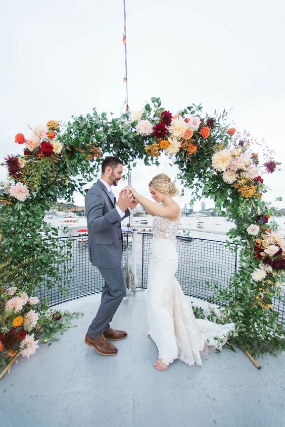 a curved wedding arch decorated with greenery, blush, orange, red and burgundy dahlias is a cooland fun idea to rock