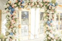 a dreamy wedding arch covered with greenery, blush roses, blue and whiote hydrangeas and pink and blush dahlias