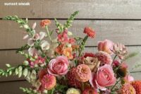 a gorgeous sunset-inspired wedding centerpiece of a gold vase, pink, orange and peachy dahlias and roses, greenery and fillers