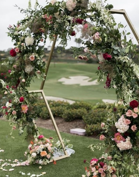 a hexagon wedding arch decorated with lush greenery, pink, peachy, orange and white flowers all over it