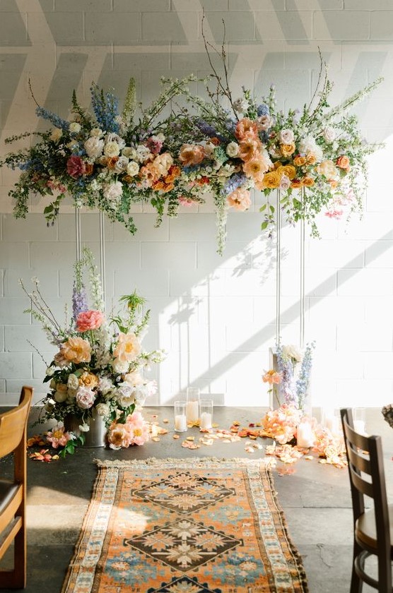 a lovely pastel wedding arch with white, orange, rust, blue and pink blooms, blooming branches and greenery and candles on the floor for a spring wedding