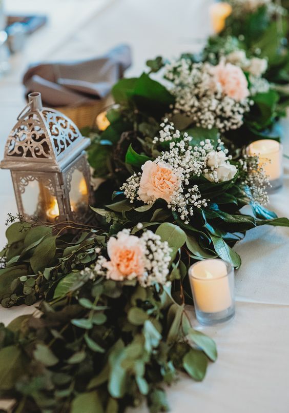 a lovely wedding table runner of greenery, blush carnations and baby's breath plus some candle lanterns is a cool idea