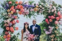 a lush and beautiful colorful wedding arch with greenery, blush, coral, purple and lilac blooms and branches and a sea view is wow