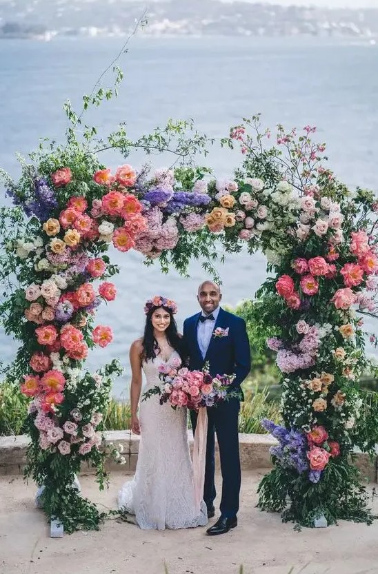 a lush and beautiful colorful wedding arch with greenery, blush, coral, purple and lilac blooms and branches and a sea view is wow