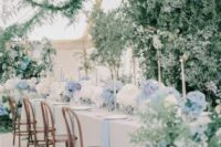 a lush and elegant blue and white hydrangea wedding table runner with some greenery and tall and thin candles is chic