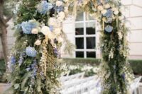 a lush and textural wedding arch done with various types of greenery, white and blush roses and blue hydrangeas looks amazing