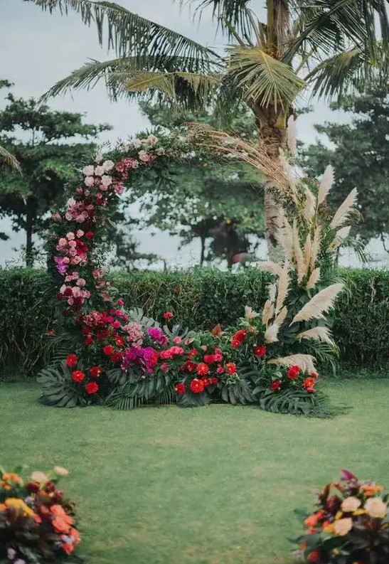 a lush round wedding arch with pampas grass, pink, blush, red and fuchsia blooms and tropical leaves