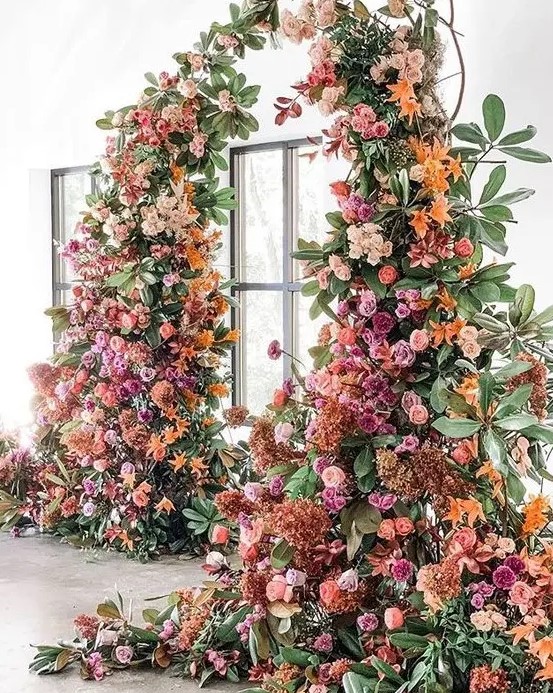 A luxurious colorful wedding arch decorated with greenery, blush, hot pink, orange and rust colored blooms is wow