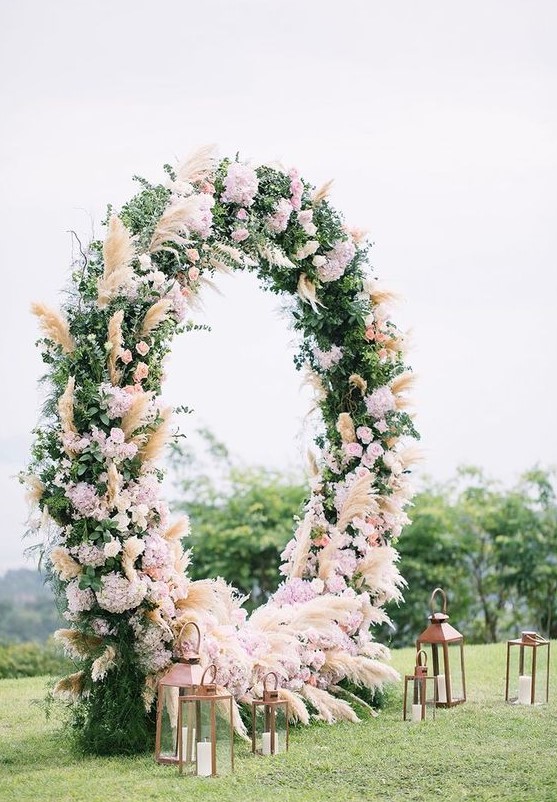 a luxurious round wedding arch with greenery, pampas grass and pink and blush blooms and candle lanterns is chic