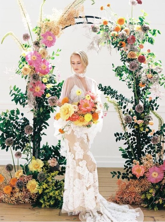 a modern refined wedding arch covered with greenery, with bold yellow, pink, orange and lilac blooms and herbs on top catches an eye