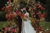 a moody fall wedding arch done with greenery and yellow leaves, with blush, coral and burgundy blooms looks very decadent and beautiful