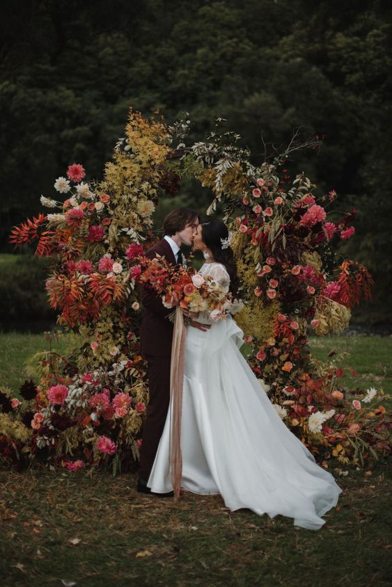 a moody fall wedding arch done with greenery and yellow leaves, with blush, coral and burgundy blooms looks very decadent and beautiful