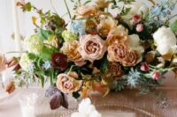 a muted color wedding centerpiece with blush roses and carnations, white, burgundy and blue blooms and greenery plus some textured foliage