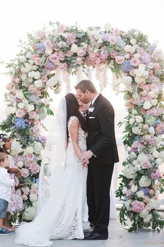 a pastel wedding arch decorated with greenery and leaves, with white and pink roses, blue and lilac hydrangeas is very lush and chic