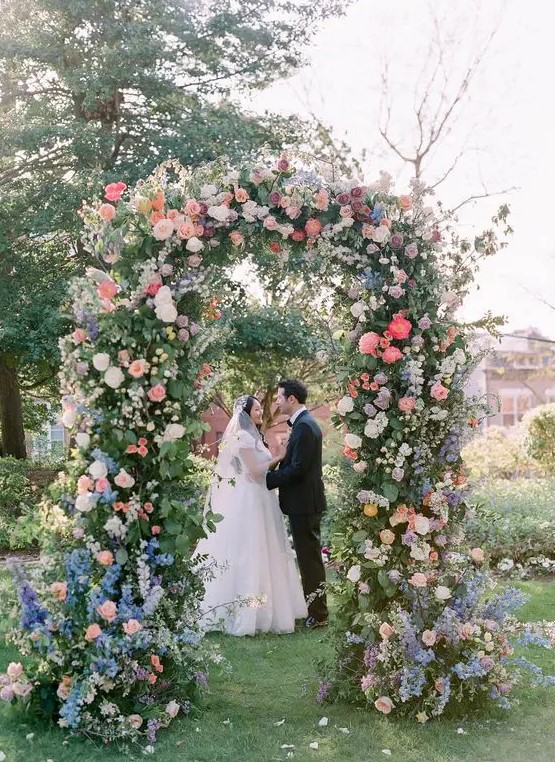 a pastel wedding arch with lots of textural greenery, blush, peachy and bold pink blooms, blue and purple ones is a cool idea