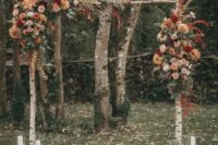 a pretty and bright rustic fall wedding arch of birch branches, rust, blush, burgundy, orange and red blooms, some eucalyptus, candle lanterns