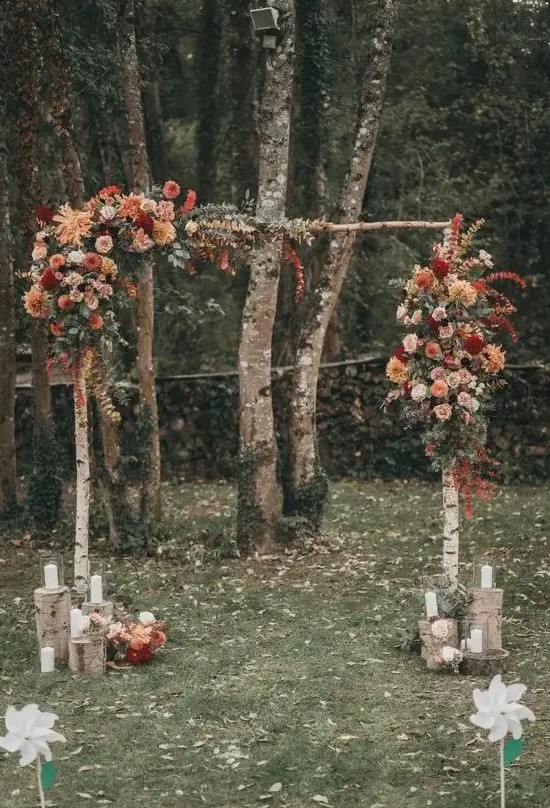 a pretty and bright rustic fall wedding arch of birch branches, rust, blush, burgundy, orange and red blooms, some eucalyptus, candle lanterns