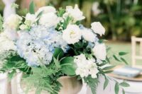 a pretty textured wedding centerpiece of white roses and blue hydrangeas, leaves and fern in a brass bow is wow