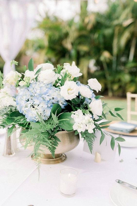a pretty textured wedding centerpiece of white roses and blue hydrangeas, leaves and fern in a brass bow is wow