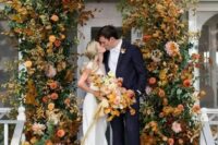 a refined and cool modern fall wedding arch composed of bold rust, yellow, mustard blooms and greenery and bright fall leaves