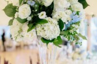 a refined tall wedding centerpiece of white hydrangeas and roses, greenery and blue fillers for a spring or summer wedding