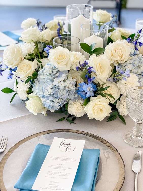 a romantic wedding centerpiece of blue hydrangeas, white roses and carnations, some fillers and pillar candles in the center