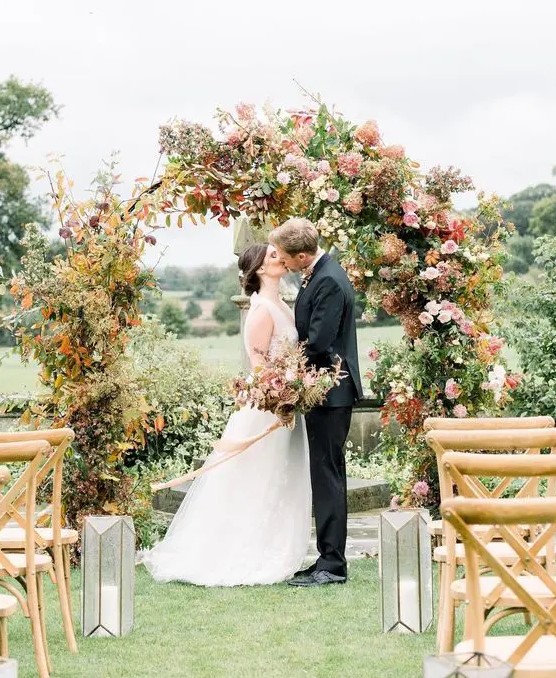 a round wedding arch with greenery and bold fall leaves, blush and pink blooms and some branches is a cool idea for a fall wedding