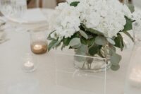a simple modern wedding centerpiece of white hydrangeas and greenery in a clear vase, with a clear acrylic table number