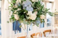 a sophisticated tall wedding centerpiece of blue hydrangeas, white roses and lots of greenery plus candles around