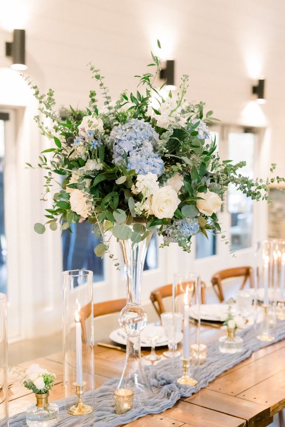 a sophisticated tall wedding centerpiece of blue hydrangeas, white roses and lots of greenery plus candles around