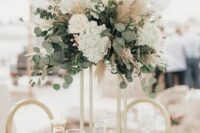 a tall boho wedding centerpiece of white roses and hydrangeas, greenery and pampas grass plus candles at the base