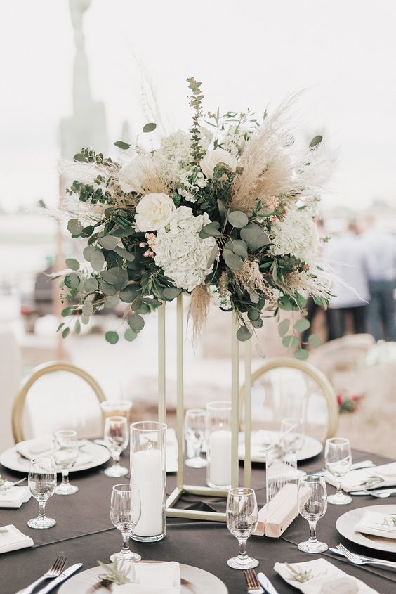 a tall boho wedding centerpiece of white roses and hydrangeas, greenery and pampas grass plus candles at the base