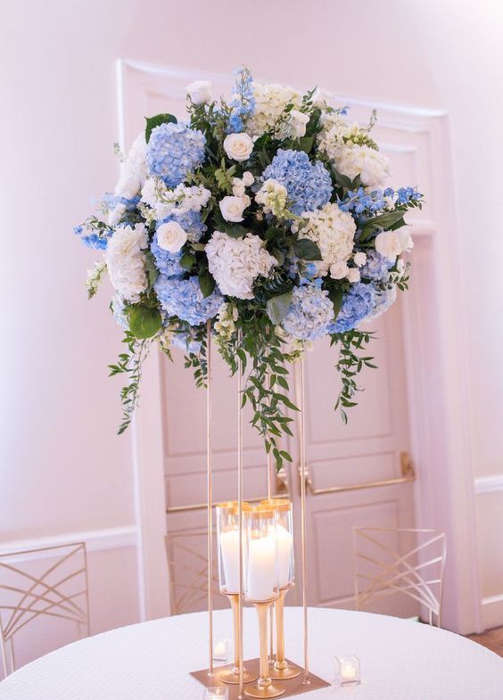 a tall wedding centerpiece of blue and white hdyrangeas, white roses and greenery and candles at the base is cool and catchy