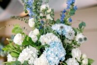 a tall wedding centerpiece of greenery, blue and white hydrangeas, white roses and bold blue fillers