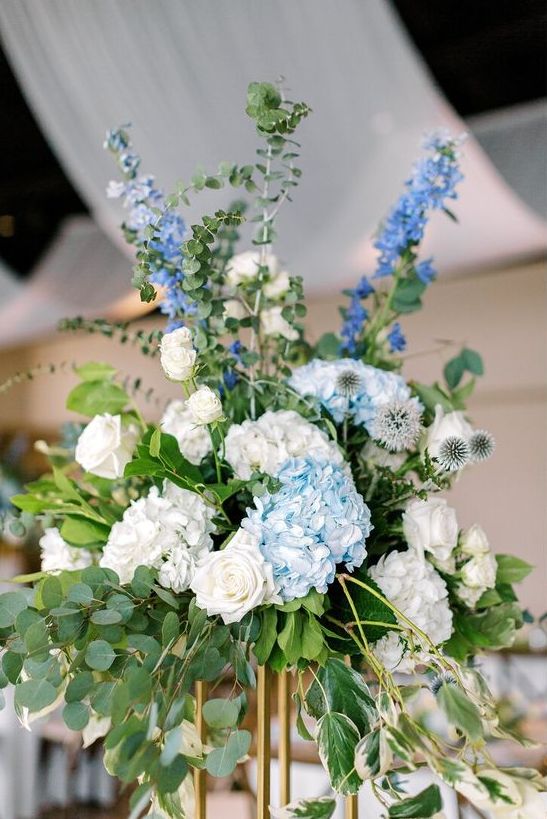 a tall wedding centerpiece of greenery, blue and white hydrangeas, white roses and bold blue fillers