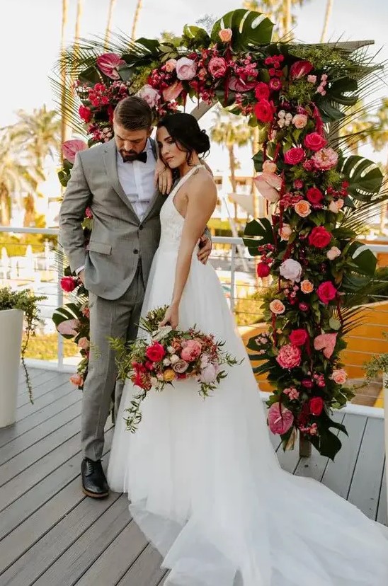 a tropical wedding arch decorated with tropical leaves, red, blush, orange and fuchsia blooms