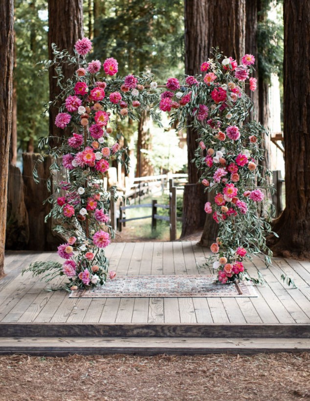 a wedding arch done with lush greenery and bold dahlias in fuchsia and pink plus deep reds