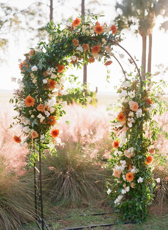 an elegant fall wedding arch decorated with greenery, white and orange dahlias is a lovely idea for a summer to fall or fall wedding