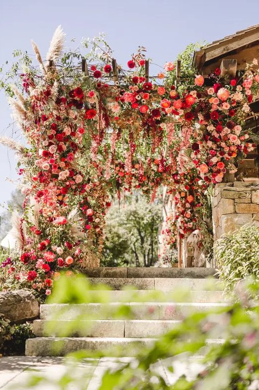 an epic wedding arbor decorated with greenery, pampas grass, pink, red and burgundy blooms is a stunning color statement