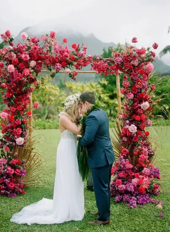 an extremely lush and colorful floral wedding arch covered with red, pink, purple blooms and some gilded leaves