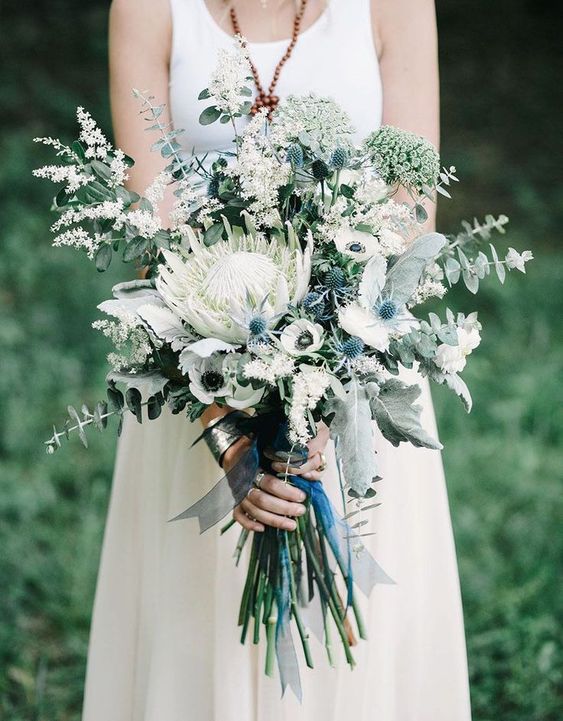 an eye-catchy wedding bouquet of white blooms, blue thistles, a large king protea and some eucalyptus is wow