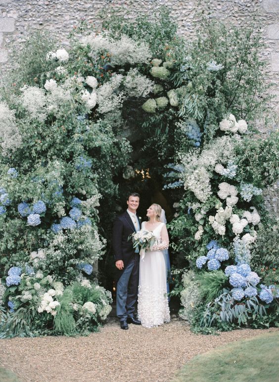 an oversized floral wedding arch done with a lot of greenery, white blooms and white and blue hydrangeas is a gorgeous idea for a spring or summer wedding