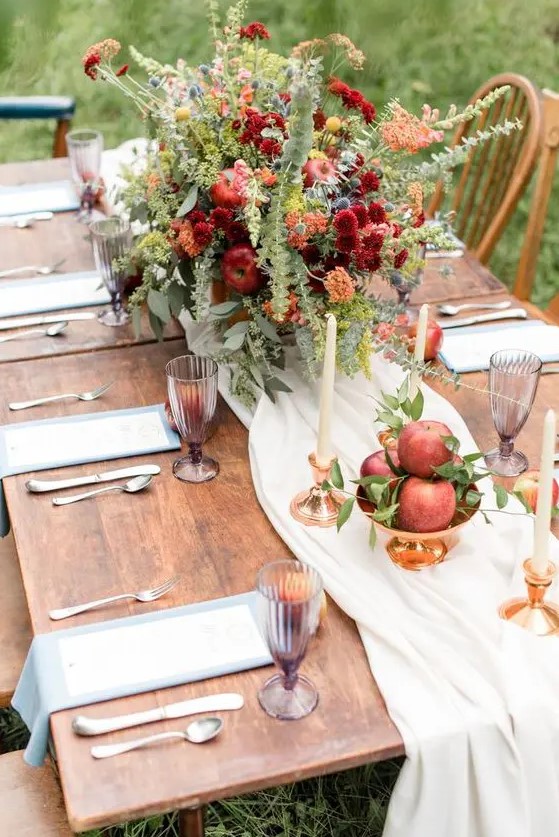 a beautiful apple orchard wedding with a neutral runner, a bold centerpiece of orange, burgundy and yellow blooms, greenery and thistles, apples and tall and thin candles