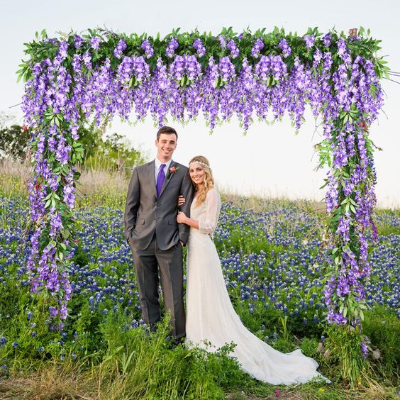 a bright wedding arch right in a blooming field, with wisteria and greenery hanging down is a cool idea for a boho wedding