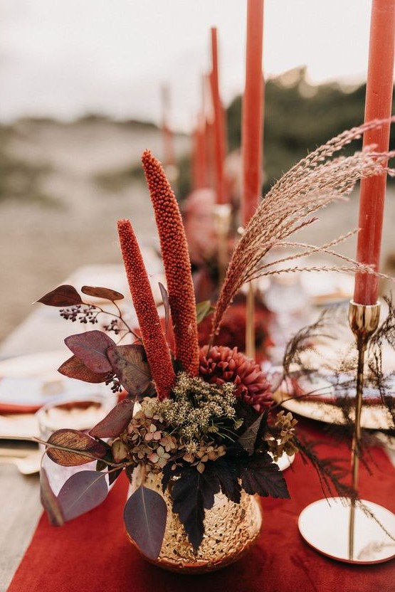 a bold fall wedding centerpiece in a gold vase, with burgundy blooms, greenery and dark foliage plus red candles