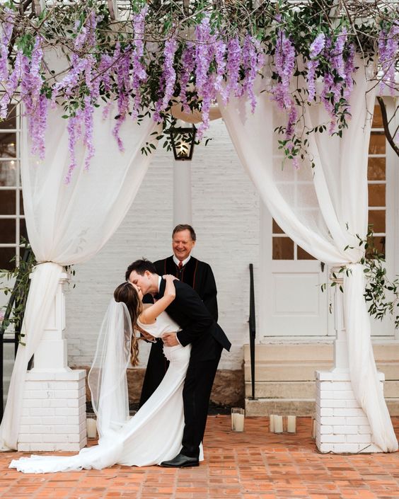 a classic wedding arch done with white curtains, wisteria and greenery plus twigs is a cool idea for a classic wedding