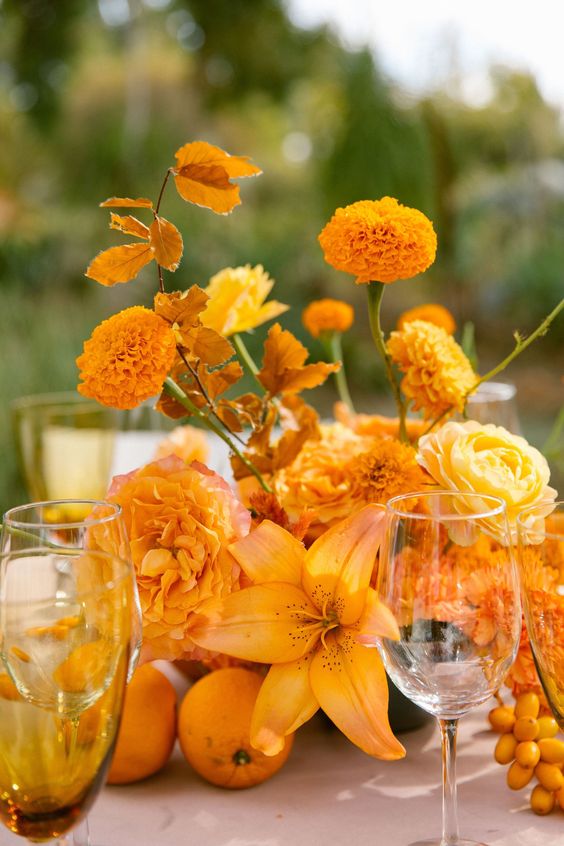 a bright fall wedding centerpiece of marigolds, roses and a lily plus matching fall leaves and citrus on the table