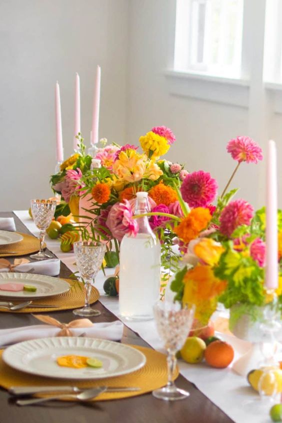 a bright wedding tablescape with bold blooms in light pink, fuchsia and yellow, marigolds, greenery, light pink candles, yellow placemats and crystal glasses